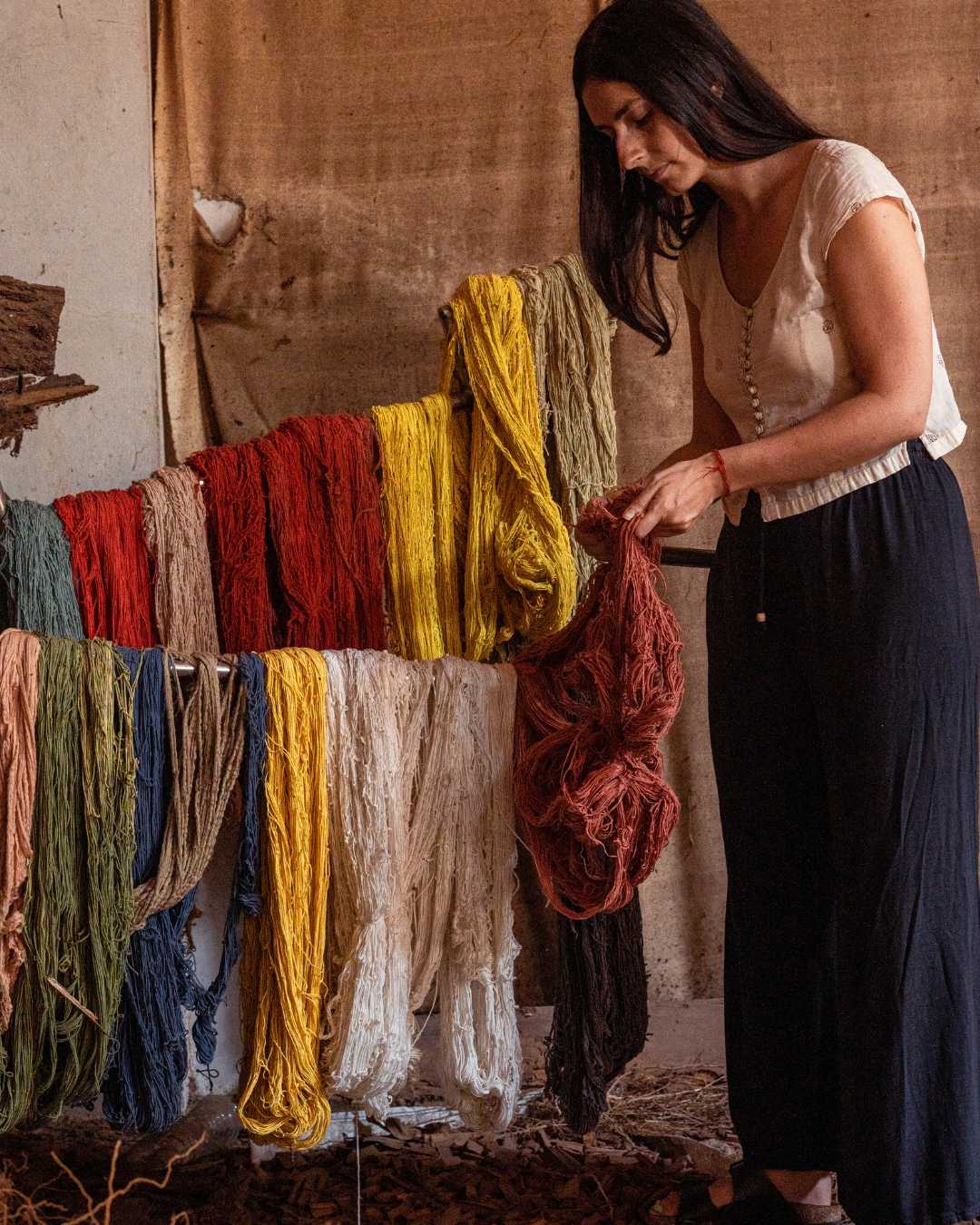 A woman in a beige top and dark skirt stands beside a display of colourful naturally dyed yarns hanging on a stand